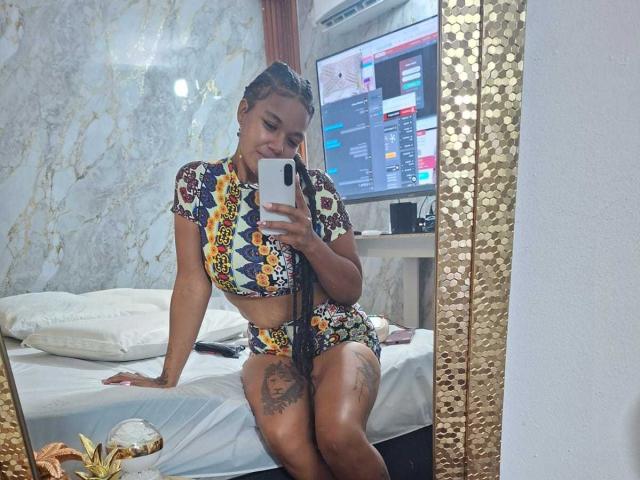 NaturaleBonyy in colorful patterned dress, holding phone, sitting on bed with legs crossed, blue and white sneakers, marble wall, TV screen, gold textured mirror, pineapple decor.