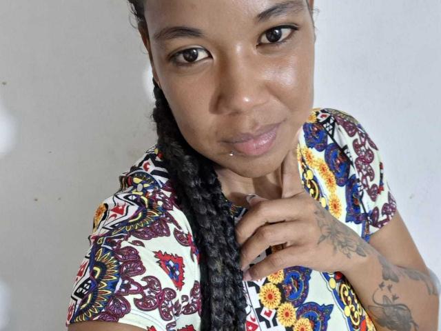 NaturaleBonyy with braided hair, brown eyes, and a subtle smile, wearing a colorful patterned shirt, showing tattoos on her hand, against a plain white background.