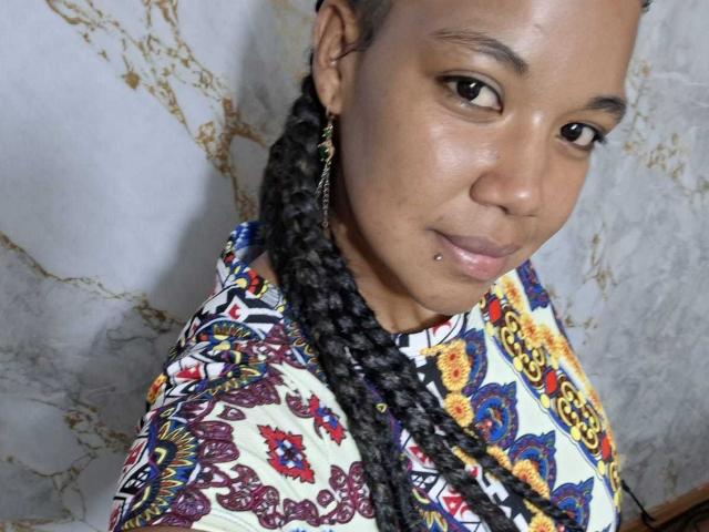 NaturaleBonyy has dark brown skin, braided black hair, a small lip piercing, and wears a colorful patterned shirt with her tits and ass not visible.