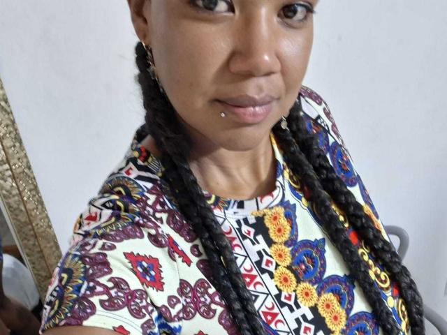 NaturaleBonyy with dark skin, braided hair, small lip piercing, and wearing a colorful, patterned dress. She's looking at the camera with a subtle smile.