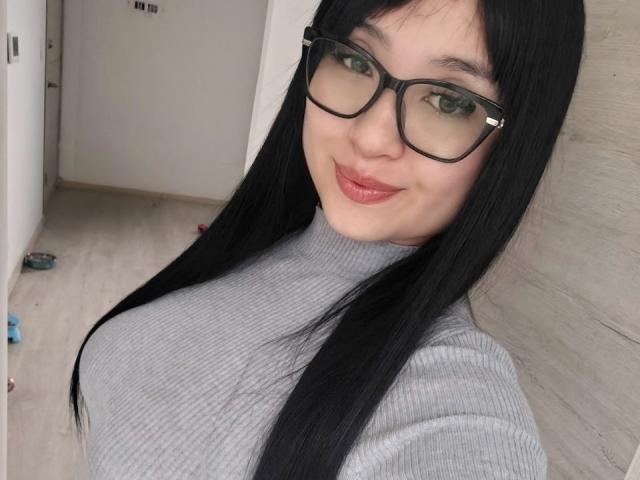 LunaBellaHotty has long, straight black hair and wears large black-framed glasses. She has light skin and is smiling with red lipstick. She is wearing a gray ribbed turtleneck that highlights her medium-sized breasts. The background is a simple, indoor setting with a wooden floor and a white door. The image focuses on her upper body and face. Her green eyes are visible through the glasses, and her hair is slightly parted in the middle. The composition is a close-up selfie.