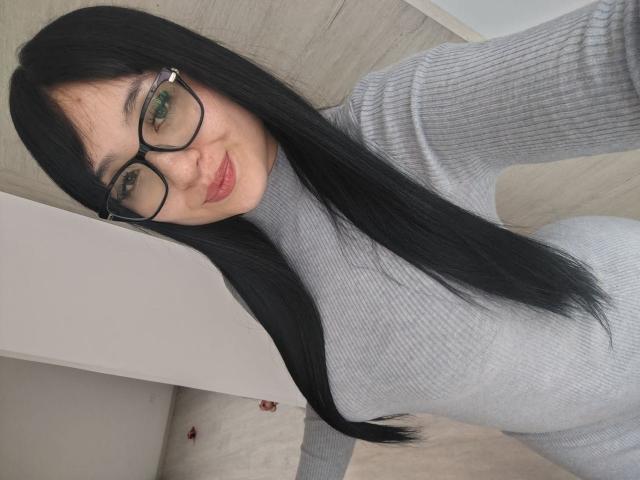 LunaBellaHotty has long black hair, green eyes, and wears black-rimmed glasses. She's smiling slightly with pink lipstick. She's dressed in a gray rib-knit turtleneck. The image is a selfie, showing her from the chest up. Her skin is light, and she's positioned slightly to the right with a wooden wall in the background. The lighting is even, highlighting her hair and glasses. The composition is simple, focusing on her face and upper body.