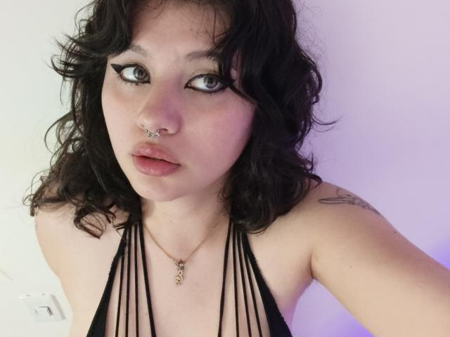 MoonBluee stands with curly black hair, red strapless top, and maroon lace panties, hand in hair, showing her curvy body and pale skin, with a confident, sultry expression.