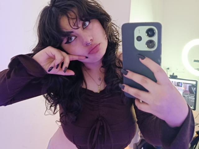 MoonBluee takes a selfie with a dark grey smartphone, holding it in her right hand and pointing at her cheek with her left. She has curly black hair, dark lipstick, and a nose ring. She wears a brown, long-sleeve top with a bow at the center. Her nails are painted black. The image is brightly lit, highlighting her pale skin. The smartphone has a large, dual-camera setup. The background is blurred.