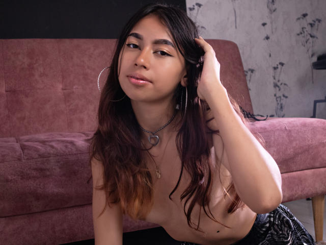ManuelaSwin, topless, sits in front of a pink velvet sofa. She has long, wavy brown hair with ombre tips, hoop earrings, and a heart-shaped necklace. Her right hand touches her hair. She has light brown skin and subtle makeup. Her small breasts are visible. She wears black, textured pants. The background has a floral-patterned wall. The image is a photograph. Her expression is slightly pouty. The overall composition is casual and intimate.