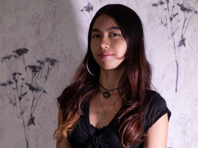 ManuelaSwin stands against a floral-patterned wall. She has long, wavy brown hair with darker roots. She wears a black, short-sleeved top with a tied knot at the chest, large silver hoop earrings, and two necklaces—one with a small circular pendant and another with a tiny, dangling figure. Her skin is smooth, and she has a subtle, natural makeup look. The lighting is soft, highlighting her relaxed expression and subtle smile. Her brown eyes are slightly downcast, adding a sense of calm to the image.