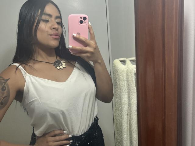 StellaBeckker takes a selfie in a bathroom, holding a pink iPhone. She has long black hair, tan skin, and is wearing a white tank top with a black high-waisted bottom. Her left arm has a tattoo of a small bird. She has white nail polish and a large, floral necklace. The mirror reflects a white towel on a wooden holder. Her expression is relaxed, with slightly pursed lips. The image is focused on her upper body.