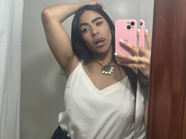 StellaBeckker takes a mirror selfie. She has medium brown skin, straight black hair, and full lips slightly parted. She wears a white, loose, sleeveless top with a black floral necklace. Her right arm is raised, hand touching her head. She holds a pink phone in her left hand. Her nails are painted white. The photo is taken in a bathroom, visible by the white curtain on the left. Her expression is neutral. The image is clear with good lighting.