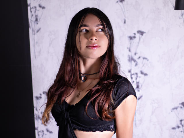 ManuelaSwin has long, straight black hair with brown tips, looking to the side with a subtle smile. She wears a black, short-sleeve, crop top with a bow tie in the front and a black choker with a small pendant. Her skin is light brown, and she has pink lipstick. The background is a white, floral-patterned wall. The photograph has a clear, well-lit focus on ManuelaSwin. The lighting is soft, highlighting her face and upper body.
