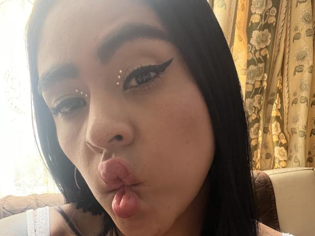 StellaBeckker's close-up face shows dark, straight hair. She puckers her lips into a kiss. Her makeup features bold black winged eyeliner with small gold studs near her eyebrows. She has a small black nose stud. Her brown eyes are slightly downcast. She wears large silver hoop earrings. The background has floral-patterned curtains. Her skin is light brown. The image is a selfie with natural lighting.