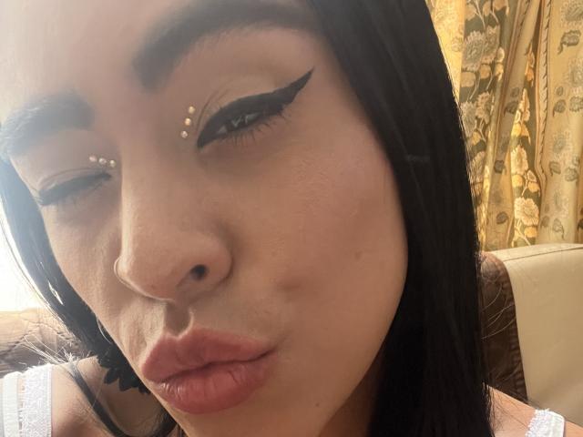 Close-up of StellaBeckker's face. She has straight black hair, medium brown skin, and is making a pouty expression. She has gold studs in her eyebrows and wears bold black winged eyeliner. Her lips are full and slightly parted. She's wearing a white lace garment, visible at the bottom. The background has floral-patterned curtains. The lighting is soft, highlighting her facial features. Her nose is slightly upturned.