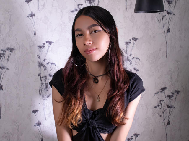 ManuelaSwin is a young woman with medium brown skin and long, wavy dark brown hair with ombre highlights. She wears a black, short-sleeve, tied-up crop top that reveals a modest cleavage. She has thin, arched eyebrows, brown eyes, and a small, relaxed smile. She wears large silver hoop earrings and a black necklace with a small pendant. The background features a grey, floral-patterned wallpaper. A black, cone-shaped lamp hangs from the top right. The composition is centered, with soft, natural light highlighting her face.