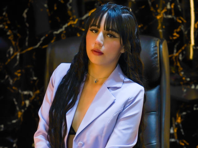 SofyBloom is a young woman with long, wavy black hair and bangs, wearing a light purple blazer with a deep V-neck. Her makeup is subtle yet effective, with pink lipstick and dark eyeliner. She has a fair complexion and a slight, contemplative expression. The blazer contrasts with the dark, marble-patterned background. Her jewelry is minimal, featuring a small pendant. The lighting is soft, highlighting her facial features and hair. The image is sharply focused on SofyBloom, with a slight blur in the background.