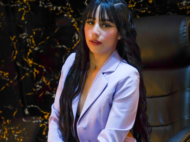 SofyBloom has long, wavy black hair with bangs, wearing a light purple blazer with a deep V-neck, revealing a hint of her cleavage. She has a light skin tone, subtle makeup, and a slight, confident smile. Her eyes are dark and she wears a delicate necklace. The background has a dark, marble-like texture with golden streaks. The image is well-lit, emphasizing her hair and blazer. Her posture is relaxed, with her hands partially visible. The blazer is slightly wrinkled, adding texture to the image.