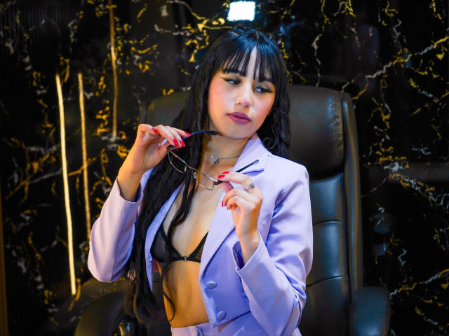 SofyBloom with long black hair and bangs, wearing a light purple blazer, black bra, and red nail polish, holds glasses in her right hand, looking slightly to the left. She has a confident expression, light makeup, and a silver necklace. The black chair and background have a glossy, marbled texture with gold streaks. The image has a sharp focus on SofyBloom and vibrant colors.