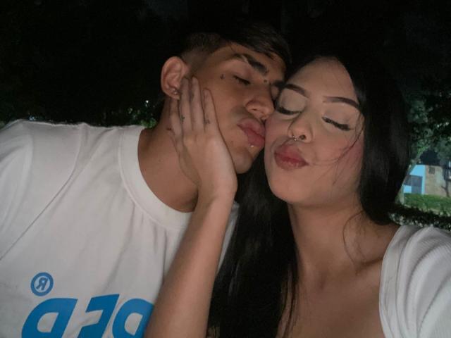 A close-up photograph of PleasurDeep, a young man with light skin and dark hair, and PleasurDeep, a young woman with light skin and long black hair. The man leans his head on the woman's hand, which rests on his cheek. He wears a white T-shirt with blue text. She has a nose ring and closed eyes, puckering her lips. The woman's hand has a small tattoo on the knuckle. The lighting is dark, emphasizing their faces. The image has a casual, intimate feel.