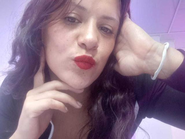 AriannaSeete is in a close-up selfie, making a puckered lip expression with bright red lipstick. She has wavy, dark brown hair and is wearing a black top. Her right hand is touching her head, and her left hand is near her chin. She has a light skin tone with freckles on her cheeks. She's wearing a white bracelet on her right wrist. The background is blurred. The image has a soft, purple-tinted light.