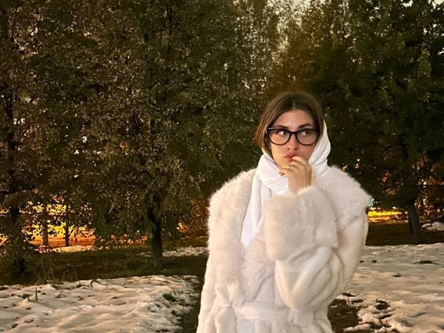 DinorahYanke in a white fluffy coat and scarf, standing in a snowy park, biting her finger, with brown hair and black glasses, looking serious.