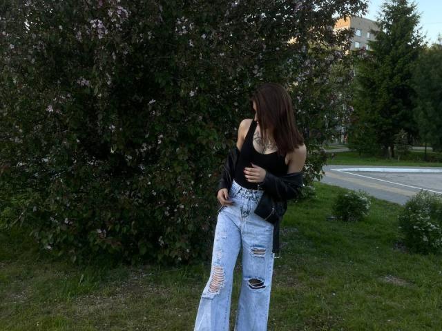 DinorahYanke with medium tits, wearing a black tank top, ripped jeans, and a black jacket, standing by a bush with purple flowers, looking to the side.