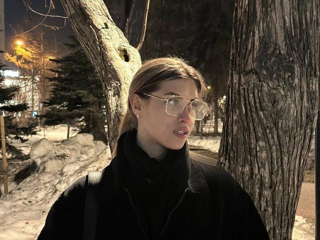 Dinorah Yanke mit Brille, schwarzem Rollkragenpullover, schwarzem Mantel und Rucksack, steht neben einem Baum in einer verschneiten, nächtlichen Umgebung. Ihr Gesäß ist nicht sichtbar..