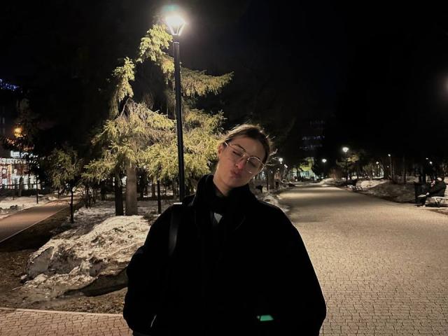 Dinorah Yanke, jaqueta preta volumosa, óculos, cabelo castanho curto, cabeça ligeiramente inclinada, noite, rua, poste de luz, neve, árvore, tijolos, sorriso discreto, alça de mochila, escuro, urbano, frio..