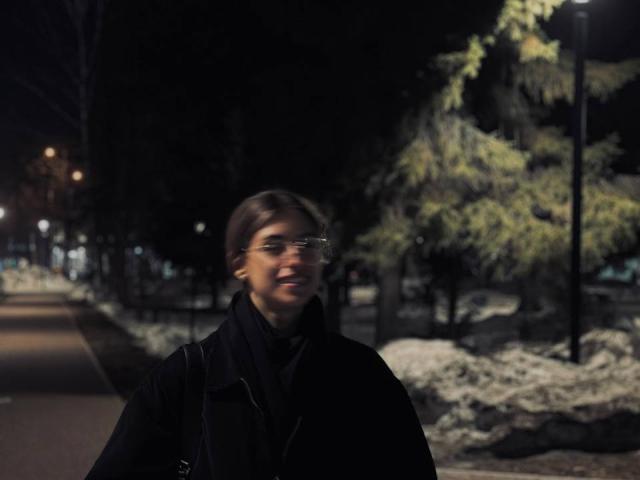 DinorahYanke with blurry face, wearing glasses and a black coat, standing in a dark, snow-covered park at night, with a streetlamp illuminating her from the side.