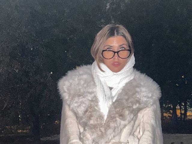 Dinorah Yanke, avec ses cheveux blonds, portant un manteau en fausse fourrure grise duveteuse, une écharpe blanche et des lunettes noires, regardant vers l'avant avec une expression neutre. Nuit, fond sombre..