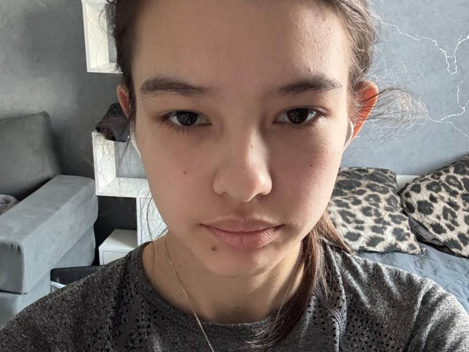 Close-up of LeathaOrmond with light brown skin and straight brown hair tied back. She has small, dark eyes, a small nose, and a slight pout. She wears a grey, textured shirt with a thin gold chain. Background includes a grey sofa, leopard print pillows, and a white staircase. The room has a grey, textured wall. LeathaOrmond's face is centered, with a neutral expression. Natural light highlights her facial features. Slight blemishes on her skin are visible. LeathaOrmond's subtle makeup emphasizes her natural look.