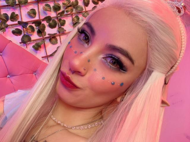 LaurenEvie6 has long, straight, platinum blonde hair, wearing a pearl hairpiece. She has dark, thick eyebrows and is making a pouty face. Her makeup includes blue star-shaped dots on her cheeks and dark, winged eyeliner. She has a silver nose ring and wears pink lipstick. Her neck is adorned with a pearl necklace. The pink, tufted background has green, vine-patterned panels. The image is brightly lit with pink and purple lighting. She has a light skin tone.