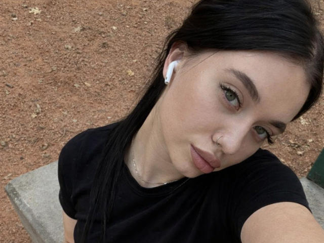 AlexaStrei has pale skin, green eyes, and black hair in a straight back style, wearing a black shirt and white earbuds, with a nose ring and subtle makeup.
