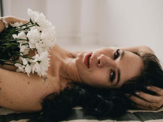 JasmineKouch with dark curly hair, lying down, holding white flowers against her bare tits, looking at the camera with red lips, soft light, subtle tattoos on her shoulder.