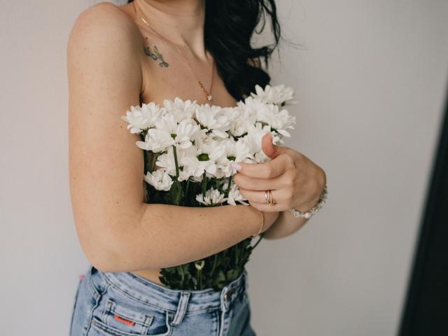JasmineKouch with white daisies in her hands, wearing a strapless top and blue jeans, has tattoos on her chest and wears a gold necklace and ring.