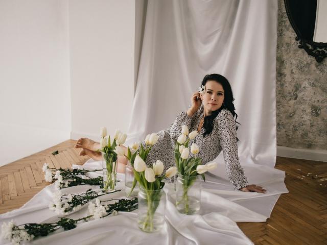 JasmineKouch with dark hair, wearing a leopard print dress, lounging on white fabric, holding a phone, surrounded by white tulips and greenery on a wooden floor.