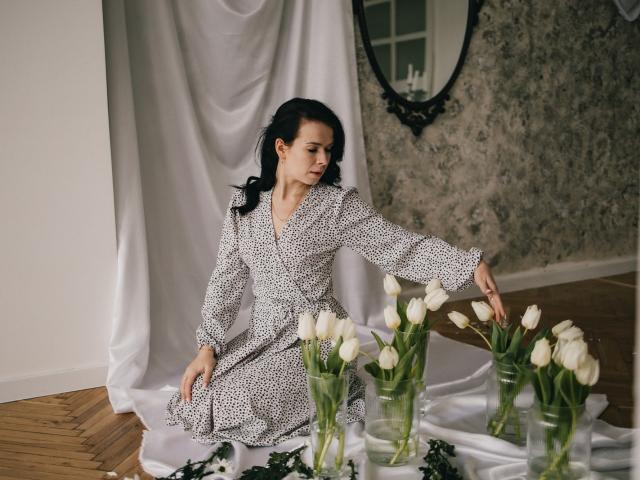 JasmineKouch with dark hair, wearing a white patterned dress, kneels, touching white tulips on a wooden floor, white fabric draped beneath, elegant mirror in the background.