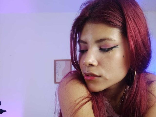 A close-up photograph of AfroditaMooreX, a young woman with red hair, closed eyes, and pink eyeshadow. She has a small nose ring on her left nostril and a visible mole under her right eye. Her right arm is bent, resting on her shoulder. She wears large hoop earrings. The background is blurred, featuring a small, framed abstract drawing. The lighting is soft, with a purple-blue hue. The image focuses on her face and upper body.