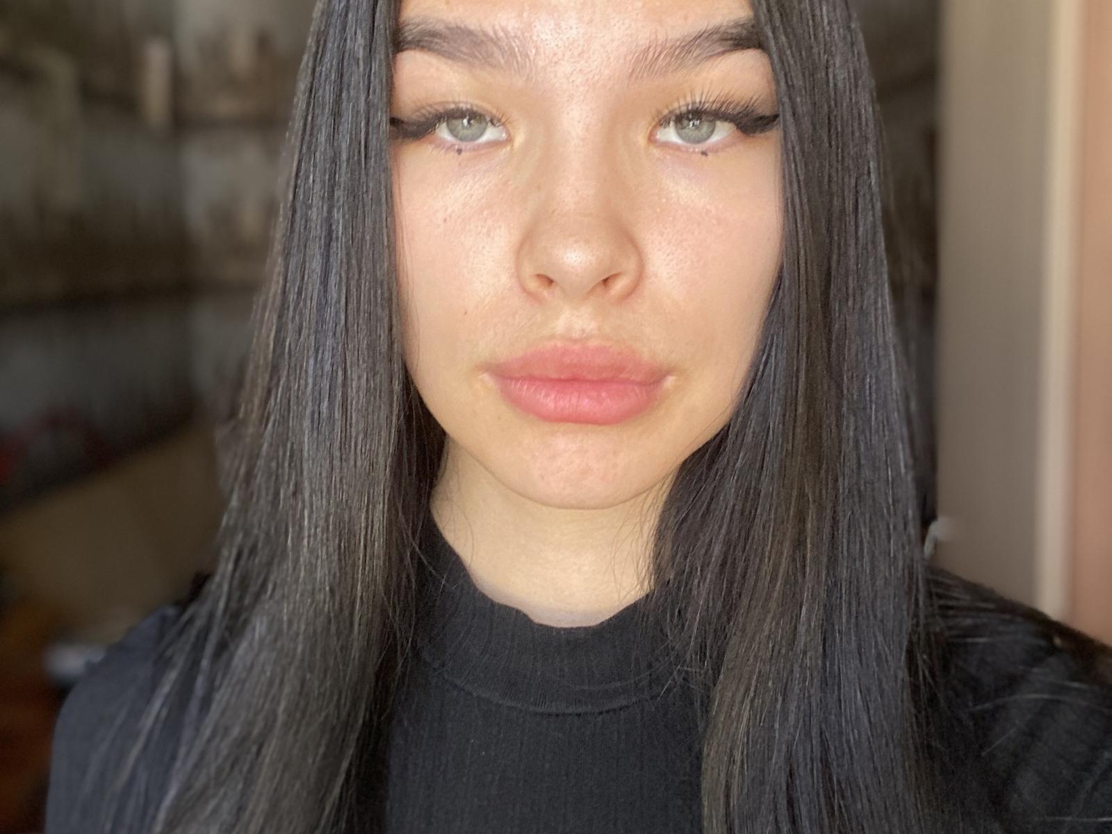 Close-up of MerriePutnam with straight, black hair framing her face. She has light green eyes, full pink lips, and a slight blush on her cheeks. Her eyebrows are thick and dark, contrasting with her fair skin. She wears a black turtleneck, and the image focuses on her face, with subtle lighting highlighting her skin texture. The background is blurred, keeping the attention on MerriePutnam's facial features and hair. The overall composition is centered, with MerriePutnam's face occupying the majority of the frame.