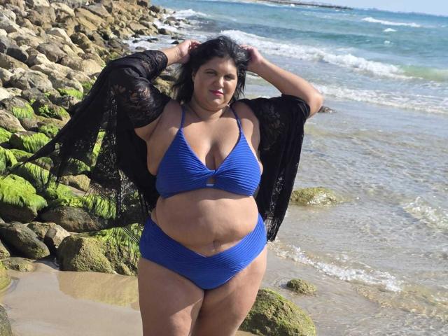 YasminQueen stands on a rocky beach, wearing a blue bikini with a halter top and high-waisted bottoms. She has a curvy, plus-size physique with visible belly rolls and large breasts. Her black, wavy hair is loose, and she has a light brown skin tone. She holds up a black, sheer, lace robe, exposing her bikini. The background shows a rocky shoreline with green moss and blue ocean waves. The sunlight highlights her body, creating a natural, relaxed vibe. Her confident, relaxed expression adds to the casual beach setting.