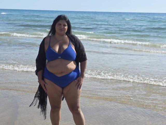 YasminQueen stands on a beach, wearing a blue bikini with a black cardigan. She has long black hair, a medium-brown skin tone, and a fuller body with noticeable belly and thigh fat. Her bikini top is slightly stretched over her large breasts. She looks directly at the camera with a neutral expression. The ocean and clear sky form the background. The wet sand under her feet reflects the sunlight.