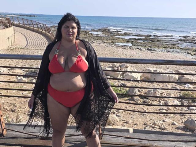 A photograph of YasminQueen standing on a wooden platform at a beach. She has a medium-brown complexion and long black hair. She's wearing a red bikini with gold ring accents and a black lace shawl. YasminQueen is plus-sized with visible rolls of fat on her midsection. The beach has a rocky shoreline with seaweed and clear blue water. Metal railings and a concrete pathway are in the background. The sky is clear and blue.