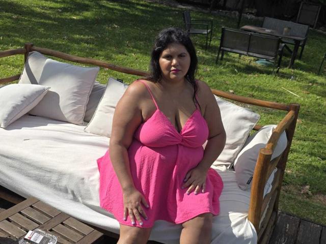 A photograph of YasminQueen, a plus-sized woman with dark, wavy hair, sitting on a wooden outdoor couch. She wears a bright pink, textured dress with thin straps and a deep V-neck, revealing ample cleavage. Her hands rest on her thighs. The couch has white cushions, and the wooden frame is visible. The green grassy background is blurred. Her expression is neutral, and she has a light brown skin tone. The image has a casual, natural style.