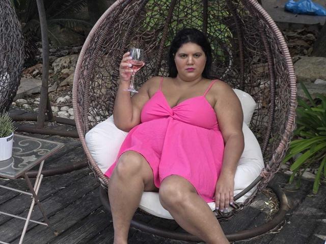 YasminQueen sits in a wicker hanging chair with a round, woven frame. She wears a bright pink, sleeveless dress with a bow at the chest. Her dark hair is straight, and she has a medium build. YasminQueen holds a glass of rosé in her right hand, her left arm resting on the chair. She has a neutral expression. The chair is cushioned with a white pillow. The wooden deck and greenery in the background are partially visible. The wicker's texture is detailed and natural.