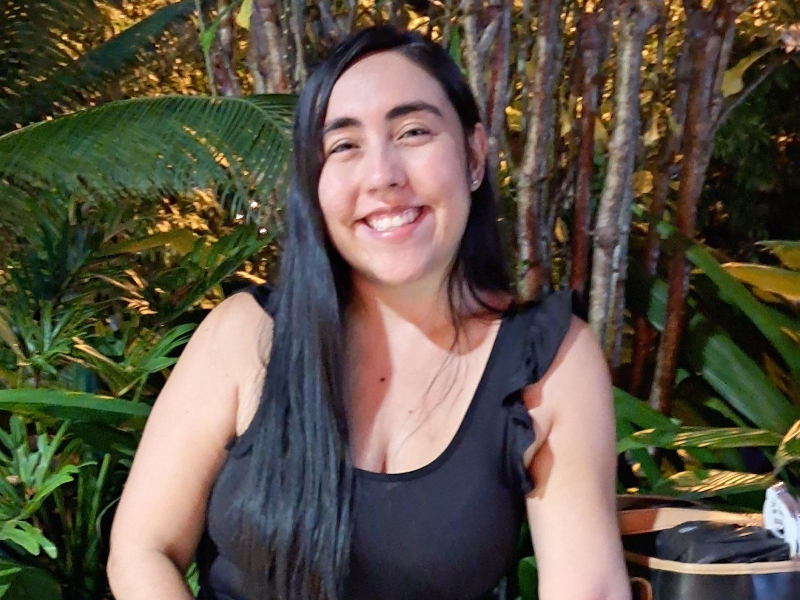 SweetyNaughty has long black hair, a smiling face with white teeth, and is wearing a black sleeveless top with ruffled edges. She has a light skin tone, dark eyebrows, and is looking directly at the camera. The top reveals a small mole on her chest. The background is lush with green tropical plants and tree trunks. The image has a casual, natural feel. She is sitting, with part of a wooden or bamboo chair visible on the right. The photo is bright with natural light.