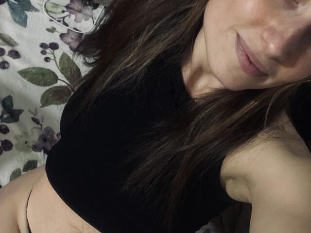 Milagres with long brown hair, black sleeveless top, and bare lower abdomen, showing a small black thong, smiling slightly, with visible pink lips and light skin.