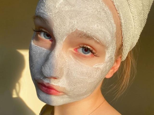 GilmaNewhouse's face is covered in white facial mask, blue eyes looking forward, small nose, pink lips, towel on head, shadow on wall, sunlight highlighting her face.