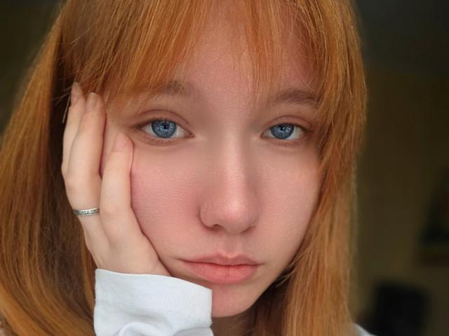 Close-up of GilmaNewhouse with fair skin and red hair, looking directly at the camera. She has blue eyes, slightly parted lips, and is resting her right hand on her cheek. She wears a simple silver ring on her right index finger. Her white shirt is visible. The image is well-lit, emphasizing her clear skin and natural red hair. Her expression is neutral.