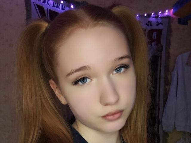 GilmaNewhouse with pale skin, blue eyes, and red pigtails, has a serious face and subtle makeup, wearing a black top, in a dimly lit room with purple string lights.