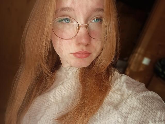 Close-up of GilmaNewhouse with fair skin, freckles, and long red hair. She wears round, gold-rimmed glasses with light green eyes. Her lips are slightly pink, and she looks directly at the camera with a neutral expression. She's wearing a white, textured sweater. The light creates a shadow pattern on her face. The composition focuses on her face and upper torso, with a blurred, brown-toned background. The image has a soft, natural light.