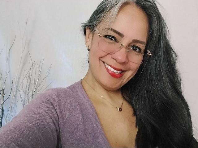 LailaCastillo with long black and silver hair, glasses, red lipstick, smiling, wearing a purple V-neck, showing her tits slightly, necklace, against a white background.