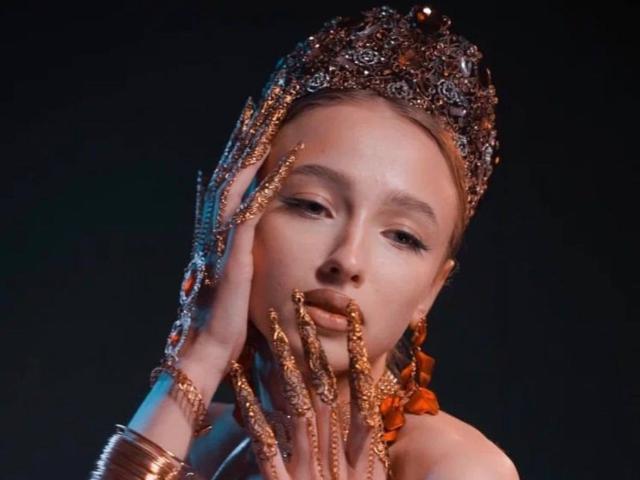 JettaMcClarty, con una elaborada y ornamentada pieza para la cabeza, sosteniendo con sus dedos dorados y joyosos sus labios, usando aretes naranjas y brazaletes dorados, sobre un fondo oscuro..