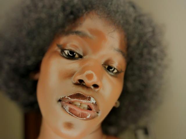 KharlaQueen mit dunkelbrauner Haut, lockigem, graubraunem Haar, glänzendem braunem Lippenstift, leicht geöffneten Lippen und einem kleinen Nasenring, die nach unten blickt und einen sanften, konzentrierten Ausdruck hat..